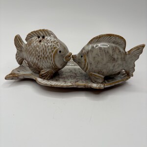 May include: A ceramic salt and pepper set featuring two fish facing each other. The fish are light brown with textured scales and fins, and sit on a matching speckled tray. The set is designed for tabletop use.