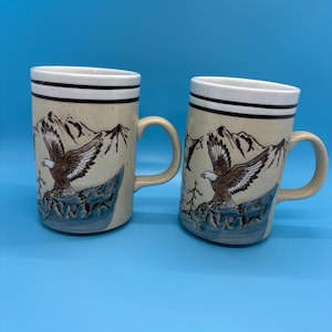 May include: Two ceramic mugs with a beige background and a mountain and eagle design. The mugs have a white rim with two black stripes and a beige handle. The eagle is brown and white, and the mountains are black.