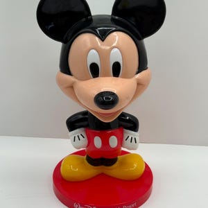 May include: A Mickey Mouse figurine with a black head and ears, a peach-colored face, and a red base. The figurine has white gloves, a red shirt with white buttons, and yellow shoes. The base reads "Walt Disney World Resort."
