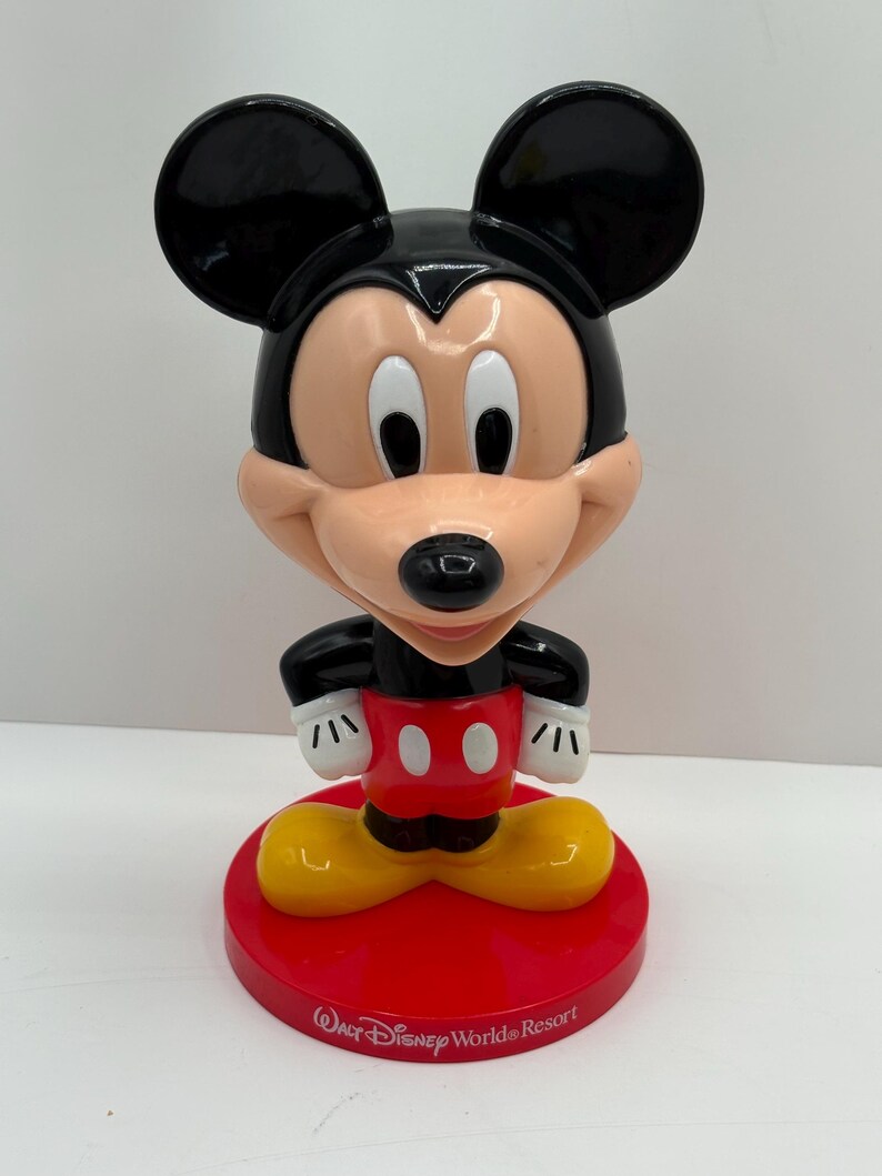 May include: A Mickey Mouse figurine with a black head and ears, a peach-colored face, and a red base. The figurine has white gloves, a red shirt with white buttons, and yellow shoes. The base reads "Walt Disney World Resort."