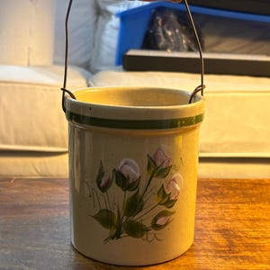 May include: A cylindrical ceramic crock with a speckled cream finish, featuring a hand-painted floral motif of pink rosebuds and green leaves. The vessel includes a dark green horizontal band near the rim and a wire handle with a wooden grip.