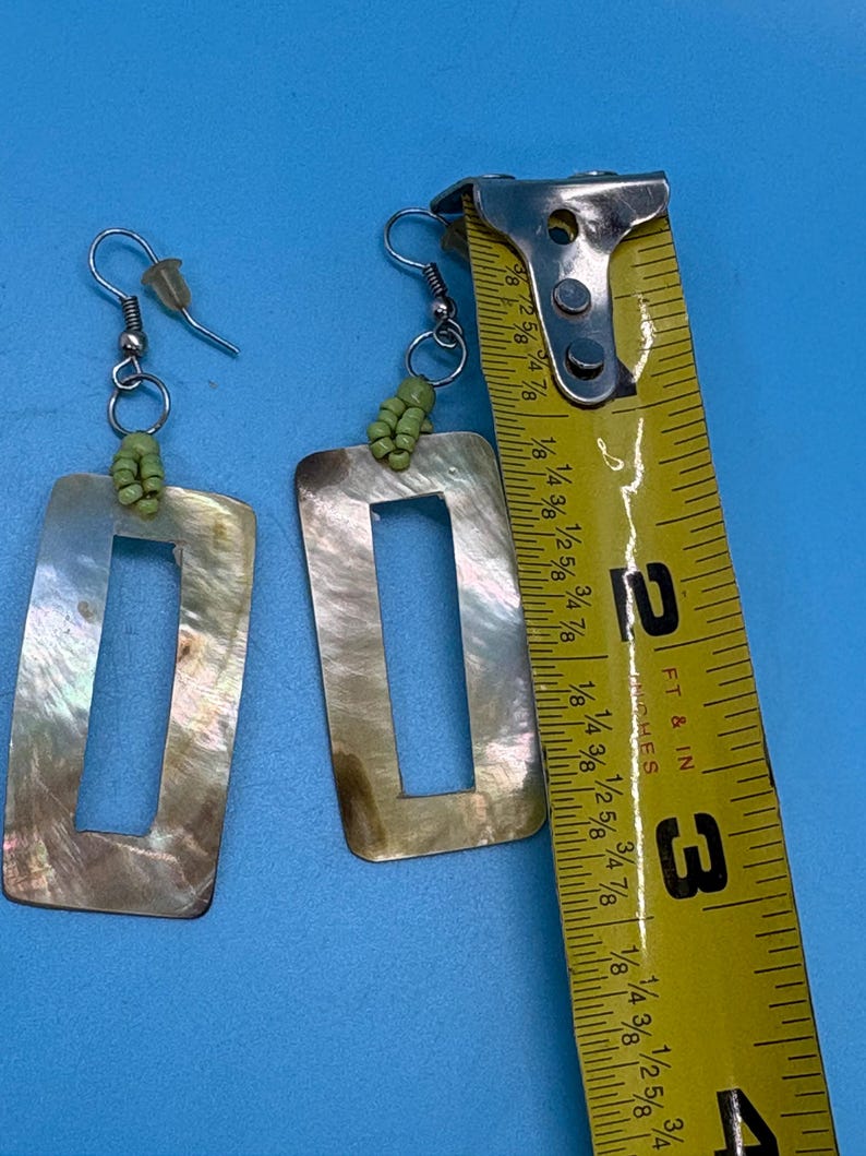 K&ouml;nnte beinhalten: Ein Paar rechteckige Ohrringe mit Perlmutt-Finish. Die Ohrringe haben ein ausgeschnittenes Rechteck in der Mitte und sind mit kleinen gr&uuml;nen Perlen verziert. Die Ohrringe sind etwa 5 cm lang.