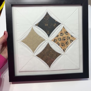 May include: Framed textile art piece featuring a geometric design. The artwork showcases four diamond-shaped fabric patches in shades of brown, tan, and olive green, set against a white background. The piece is encased in a black frame.