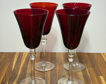 May include: Four red wine glasses with clear stems and bases are arranged on a wooden surface. The glasses have a deep red color and a classic wine glass shape. The stems have a decorative design.