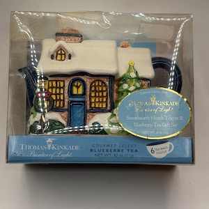 Peut inclure: Un coffret cadeau Thomas Kinkade Stonehearth Hutch avec thé à la myrtille. La théière a la forme d'un chalet enneigé avec un sapin de Noël. La boîte indique "Gourmet Select Blueberry Tea" et contient 6 sachets de thé.