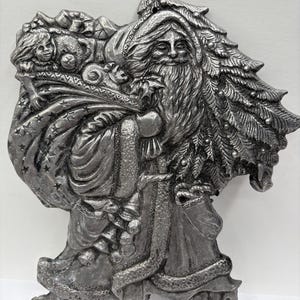 Può includere: Un ornamento natalizio in peltro argentato che raffigura Babbo Natale che porta un sacco di regali e un albero di Natale. L'ornamento è dettagliato con una superficie testurizzata e presenta una rappresentazione realistica del viso di Babbo Natale.