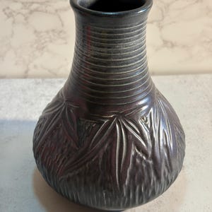 Könnte beinhalten: Eine dunkelgraue Keramikvase mit strukturierter Oberfläche. Die Vase hat einen breiten Boden mit detailliertem Blättermuster und einen gerippten Hals. Die Öffnung ist dunkel und glatt.