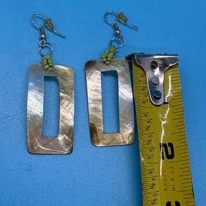 K&ouml;nnte beinhalten: Rechteckige H&auml;ngeohrringe mit Perlmutt-Finish. Die Ohrringe sind ca. 6,35 cm lang. Jeder Ohrring hat einen silberfarbenen Haken und eine kleine Gruppe gr&uuml;ner Perlen oben.