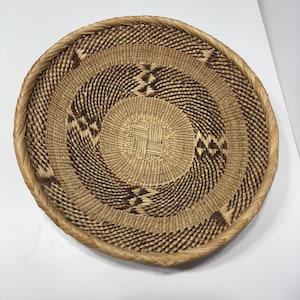 May include: A round, woven basket with a spiral design. The basket features alternating bands of light and dark brown fibers, creating a textured pattern. The rim is reinforced with a braided border. This decorative basket is suitable for home decor.