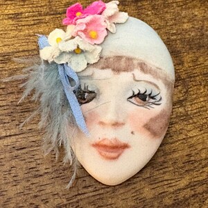 May include: A ceramic brooch shaped like a human face with painted features, including brown eyes, dark eyelashes, and pink lips. The face is adorned with a cluster of small fabric flowers in shades of pink and white, a blue ribbon, and grey feathers.