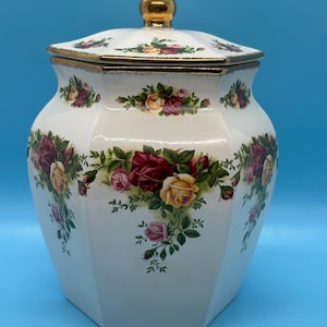 May include: A white ceramic jar with a lid, featuring a floral rose pattern in shades of red, yellow, and pink. The jar has a gold-colored knob and trim. The jar is a decorative kitchen storage container.