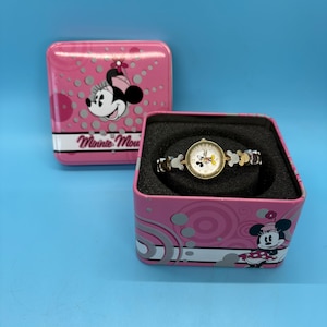 May include: A pink Minnie Mouse watch in a matching pink tin. The watch has a white face with Minnie Mouse graphics and a silver band. The tin features Minnie Mouse graphics and the words "Minnie Mouse".