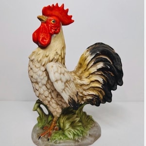 Peut inclure: Une figurine de coq en céramique avec une crête et des barbillons rouge vif, des plumes couleur crème avec des accents bruns et une queue noire foncée. L'oiseau repose sur une base sculptée d'herbe verte et de pierre grise, aux détails soignés.