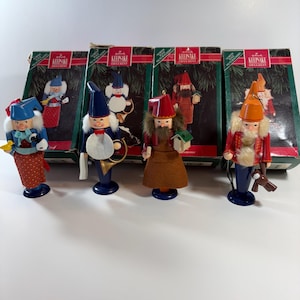 May include: A collection of Hallmark Keepsake ornaments featuring wooden figures of gnomes and other characters. The figures are painted in various colors, including blue, red, and brown, and are displayed in front of their original boxes.