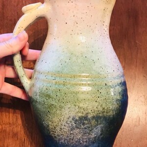 Può includere: Brocca in ceramica fatta a mano con un design sfumato maculato. I colori passano dal crema al verde e poi a un blu intenso alla base. Presenta un manico curvo e un beccuccio per versare.