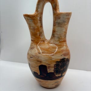 Może przedstawiać: Ceramiczny wazon z unikalnym uchwytem, ​​przedstawiający pustynny krajobraz. Wazon ma jasnobrązową i beżową kolorystykę z namalowaną sceną kaktusów, drzewa i ptaków. Wazon ma około 25 cm wysokości.