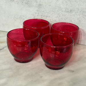 May include: Four small, round, ruby-red glass tumblers. The glasses have a simple, elegant design with a slightly flared rim. The glasses are made of a transparent material, allowing light to pass through and reflect off the surface.