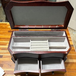May include: A dark wood jewelry box with a hinged lid, a gray velvet-lined interior, and two pull-out drawers. The lid features small metal hooks, while the main compartment includes a ring holder and open storage sections for organizing items.