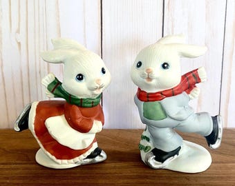 May include: Two ceramic bunny figurines, one in a red dress and green scarf, the other in a gray outfit and red scarf, both ice skating. The bunnies have white fur, blue eyes, and black ice skates. The figurines are on a wooden surface.