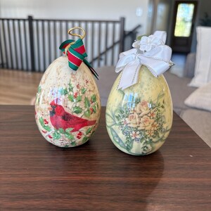 May include: Two decorative egg-shaped ornaments. One features a red cardinal bird and holly on a cream background, with a red and green ribbon. The other has a floral design with a white bow.