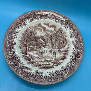 May include: A circular ceramic plate featuring a brown and white winter landscape scene. The central image shows a house, trees, and figures ice skating. The rim is decorated with a floral and wheat pattern in brown.