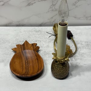 May include: A wooden pineapple-shaped bowl and a decorative candle lamp. The lamp has a brass pineapple base, a cream-coloured candle sleeve, and a clear, flame-shaped bulb. The bowl is made of wood with a natural finish.