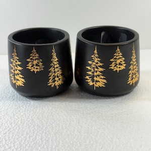 Könnte beinhalten: Zwei schwarze Kerzenhalter mit goldenen Kieferndesign. Die Halter haben eine abgerundete Form und eine glatte, matte Oberfläche. Die goldenen Bäume sind detailliert und verleihen dem Design einen Hauch von von der Natur inspirierten Eleganz.