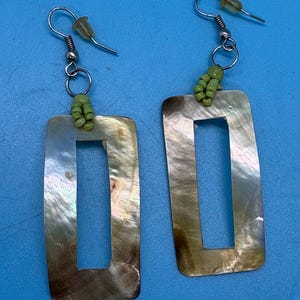 K&ouml;nnte beinhalten: Ein Paar rechteckige Ohrringe mit Perlmutt-Finish. Jeder Ohrring hat einen silberfarbenen Haken und eine Ansammlung kleiner gr&uuml;ner Perlen. Die Ohrringe haben ein hohles, offenes Rechteck-Design.