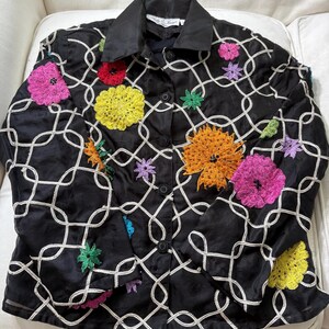 May include: Black button-down jacket with a white geometric pattern and colorful embroidered flowers. The jacket features a black collar and buttons, with vibrant pink, yellow, orange, and green floral accents. A unique, eye-catching piece.