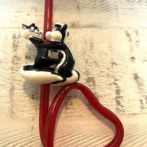 Puede incluir: Una pajita roja en forma de corazón con una decoración superior que presenta dos mofetas de dibujos animados en un abrazo amoroso. Las mofetas son negras y blancas con una base blanca. La pajita está hecha de un material rojo transparente.