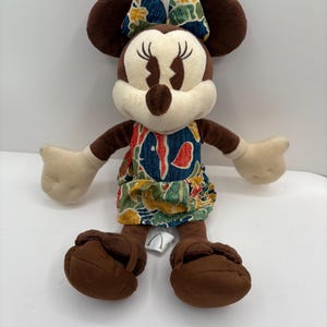 May include: A plush Minnie Mouse doll with brown ears, arms, and feet. She wears a dress and bow with a colorful floral pattern. The doll has a cream-colored face and hands, and is sitting upright.