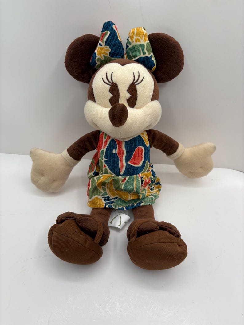 May include: A plush Minnie Mouse doll with brown ears, arms, and feet. She wears a dress and bow with a colorful floral pattern. The doll has a cream-colored face and hands, and is sitting upright.