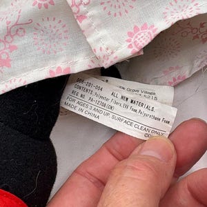 May include: A close-up of a black stuffed animal with a red accent. A tag is visible with text including "ALL NEW MATERIALS" and "MADE IN CHINA". The background includes a white fabric with a pink floral pattern.