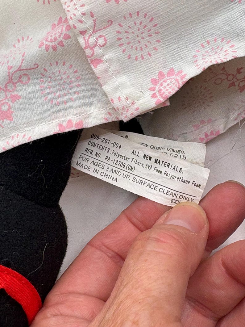 May include: A close-up of a black stuffed animal with a red accent. A tag is visible with text including "ALL NEW MATERIALS" and "MADE IN CHINA". The background includes a white fabric with a pink floral pattern.