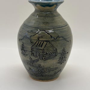 Könnte beinhalten: Eine handgefertigte Keramikvase mit rustikalem Design. Die Vase zeigt eine Hüttenszene mit Bergen und Bäumen in Blau- und Grautönen. Die Vase hat einen ausgestellten Rand und einen abgerundeten Körper, ideal für Blumen oder als dekoratives Objekt.