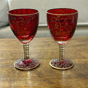 Könnte beinhalten: Zwei rote Weingläser mit goldenen Akzenten. Die Gläser haben einen goldenen Rand und ein goldenes Filigranmuster. Die Stiele sind goldfarben und die Böden haben einen goldenen Rand. Auf einer Holzoberfläche.