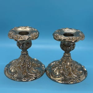 Puede incluir: Un par de candelabros de metal plateado con un intrincado patrón en relieve de uvas y hojas de parra. Cada candelabro tiene un borde festoneado ancho, un tallo central delgado y una base circular amplia con detalles ornamentados.