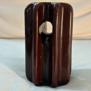 Può includere: Un isolatore elettrico cilindrico in ceramica di colore marrone scuro, caratterizzato da una finitura lucida e scanalature verticali. L'oggetto presenta un'apertura orizzontale centrale vicino alla sommità, pensata per il passaggio di cavi.