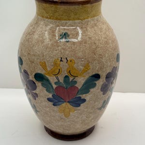 May include: A beige ceramic vase with a speckled finish, featuring hand-painted floral and bird designs. The vase has a wide opening and a brown rim and base. The artwork includes yellow birds, purple flowers, and a red heart.