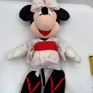 May include: A plush Minnie Mouse doll wearing a pink and white floral kimono with a red obi. The doll has black ears, a pink bow, and black shoes with red straps. The doll is smiling.
