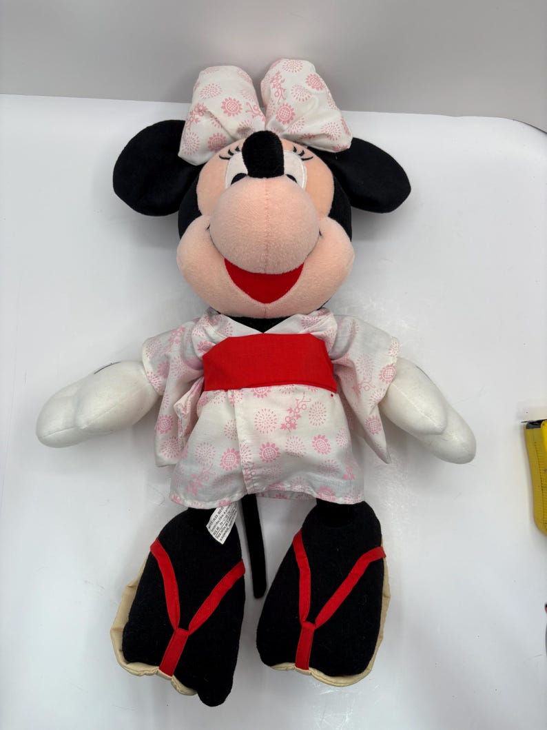 May include: A plush Minnie Mouse doll wearing a pink and white floral kimono with a red obi. The doll has black ears, a pink bow, and black shoes with red straps. The doll is smiling.