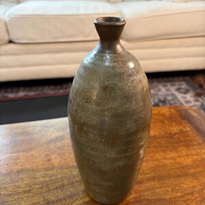 May include: A tall, slender ceramic vase with a tapered neck and a flared rim. The body of the vase features a mottled, earthy green and brown glaze with a textured, matte finish that transitions to a darker, metallic-toned glaze at the top rim.