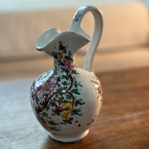 Peut inclure: Un pichet en céramique blanche orné d'un motif floral peint à la main avec des touches de violet, de jaune et de vert. Le récipient présente un corps arrondi, un bec verseur évasé et une anse incurvée partant du col.