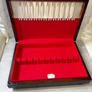 May include: A rectangular wooden storage case with a dark brown finish, featuring a hinged lid. The interior is lined with bright red fabric, including a bottom section with slots for flatware and a top section with white fabric and hooks.