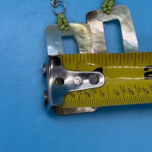 K&ouml;nnte beinhalten: Ein Paar quadratische Ohrringe aus Perlmutt mit silbernen Haken und gr&uuml;nen Perlenakzenten. Die Ohrringe sind etwa 2,5 cm lang. Die Ohrringe werden auf blauem Hintergrund pr&auml;sentiert.