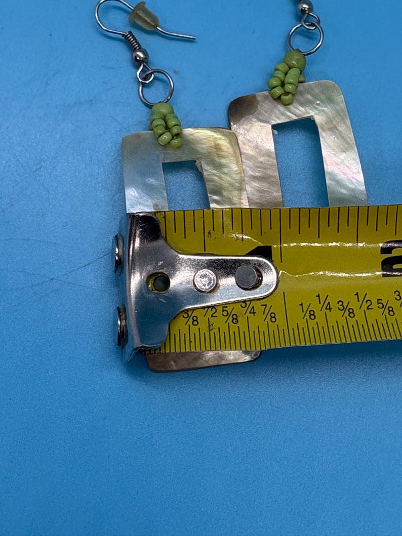K&ouml;nnte beinhalten: Ein Paar quadratische Ohrringe aus Perlmutt mit silbernen Haken und gr&uuml;nen Perlenakzenten. Die Ohrringe sind etwa 2,5 cm lang. Die Ohrringe werden auf blauem Hintergrund pr&auml;sentiert.