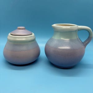 May include: A ceramic sugar bowl with a lid and a small pitcher. The sugar bowl and pitcher are in shades of blue, purple, and green. The sugar bowl is round with a lid. The pitcher has a handle and a spout. The background is a solid light blue.