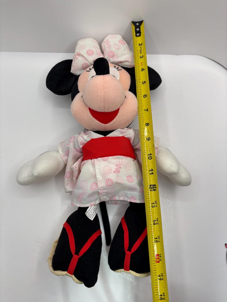 May include: A Minnie Mouse plush toy, approximately 16 inches tall, wearing a pink and white floral kimono with a red obi. The toy has black ears, a pink face, and a red bow.