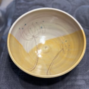 May include: A ceramic bowl with a smooth, glossy finish featuring a two-tone color scheme of cream and yellow. The interior is decorated with hand-drawn, dark brown line art of two dandelion-like floral stems with small blue dots at the tips.
