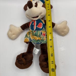 May include: A Minnie Mouse plush doll, approximately 12 inches tall, wearing a colorful dress and bow. The doll has brown ears, arms, and legs, with beige hands and face. The dress features a blue, red, and green tropical pattern. The doll is wearing brown sandals.